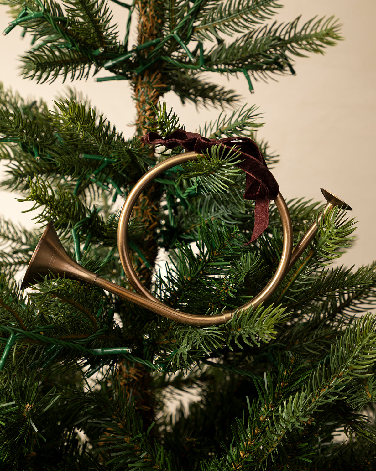 The French Horn Ornament features a brass finish and a burgundy ribbon, hanging on a green holiday tree decorated with lights, by Vendixs