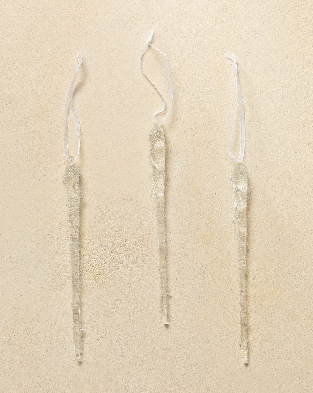 The Frosted Icicle Ornaments (Set of 3) feature clear glass icicles with white looped ribbons, displayed against a beige textured background—ideal for adding sparkle to your Christmas tree, by Vendixs