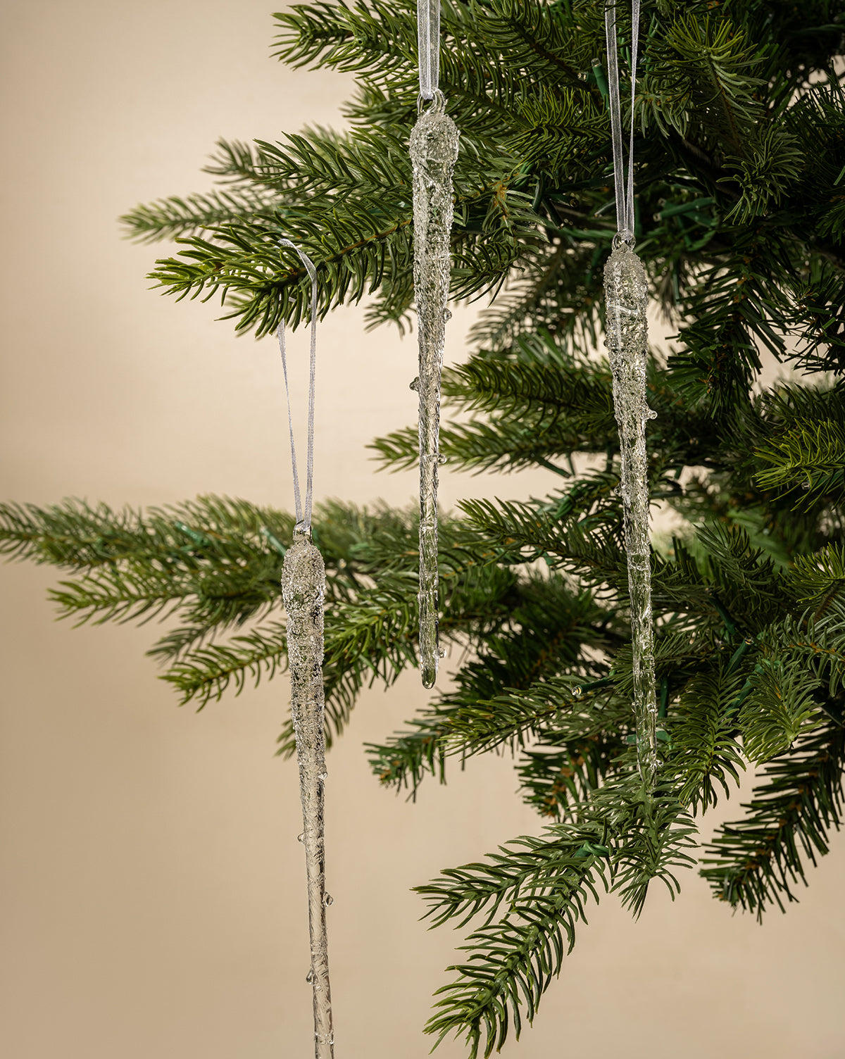 Three Frosted Icicle Ornaments (Set of 3) hang from the branches of a green artificial Christmas tree, displayed against a neutral beige background, by Vendixs