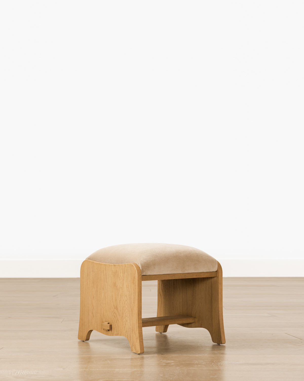 The Makerspalm Gable Ottoman features a Drift Oak frame, rounded edges, a low shelf, and a beige Fawn Velvet seat cushion. Styled simply, it stands on a wooden floor against a white wall. Vendixs