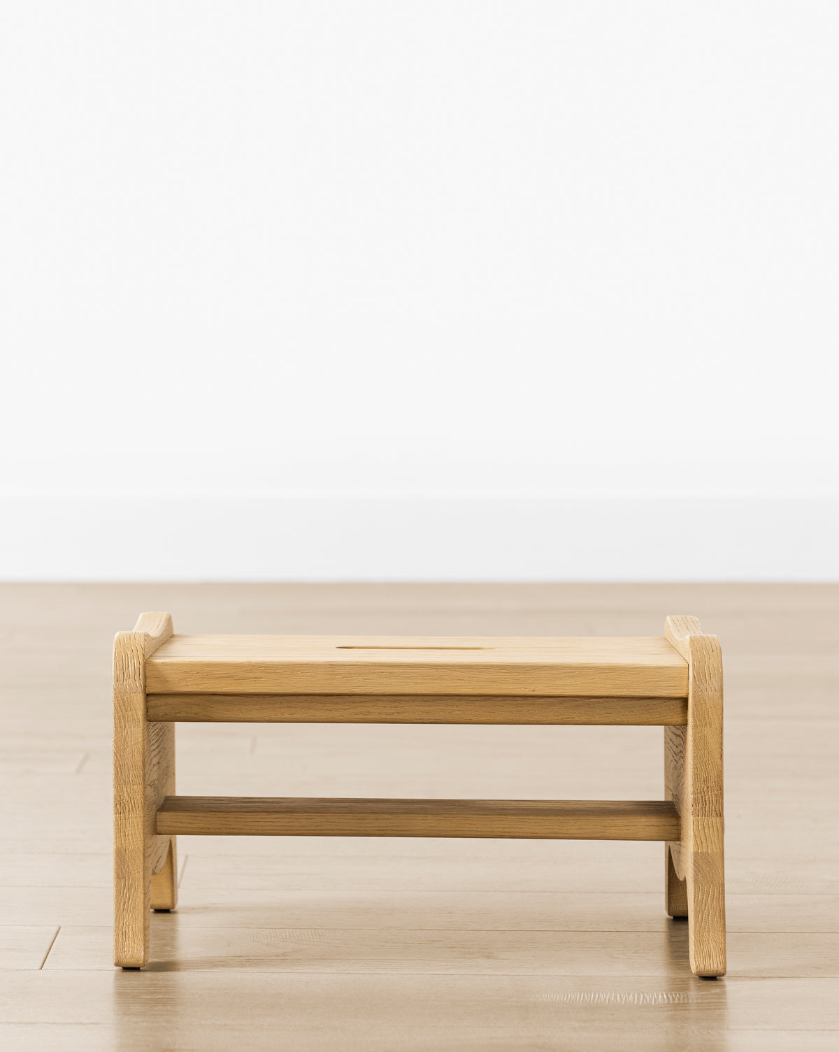 The Gable Step Stool by YANG CHENG WOODEN INDUSTRIES INTERNATIONAL (VIETNAM) CO LTD, made from drift oak with mortise and tenon joinery, features a rectangular top with rounded edges and is set on a wooden floor by a white wall. Vendixs