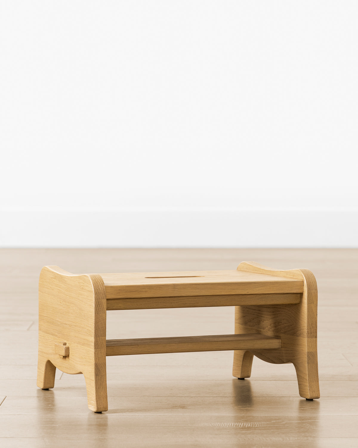 The Gable Step Stool by YANG CHENG WOODEN INDUSTRIES INTERNATIONAL (VIETNAM) CO LTD, crafted in drift oak with curved edges, a central cut-out handle, and mortise and tenon joinery, stands on a light wooden floor against a white wall. Vendixs