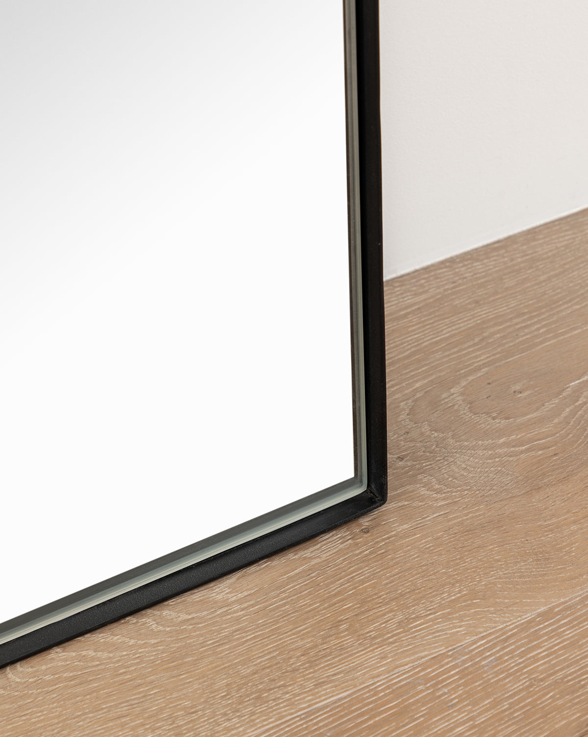 A close-up of the Gala Floor Mirror’s thin black frame resting on light wood beside a white wall—an ideal stylish addition to your bedroom mirror setup, by Vendixs