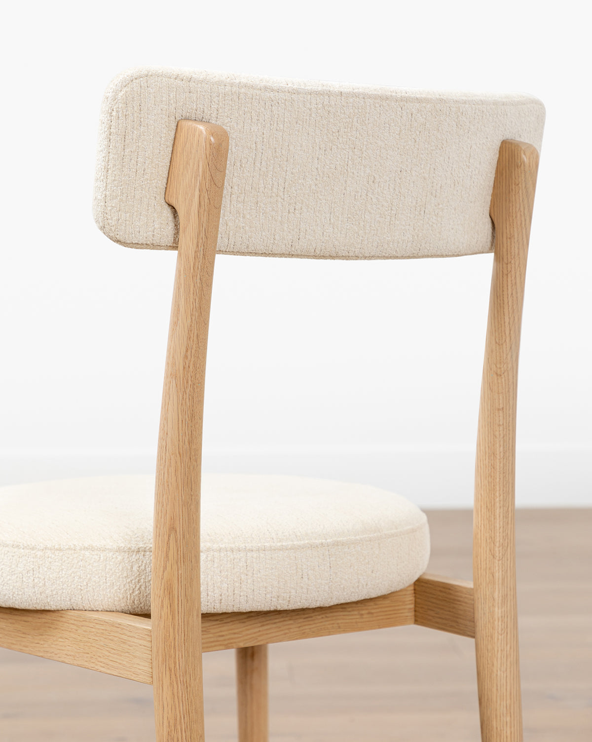 The Garance Dining Chair features a light performance velvet seat and curved backrest in white oak, shown from behind on a wooden floor against a plain white wall by Vendixs