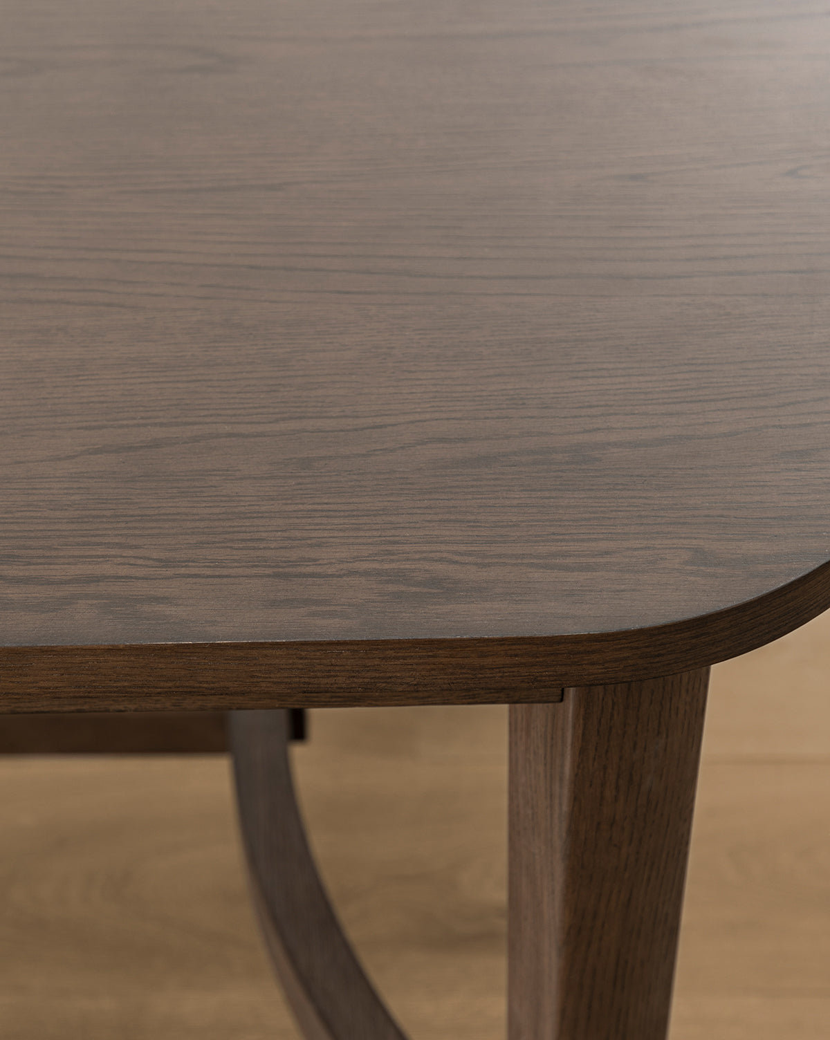 Close-up of the Gardiner Dining Table reveals a rounded corner, visible wood grain, and a single leg. The table features a smooth Walnut Oak finish, contrasting with the lighter-colored wooden floor beneath, by Vendixs