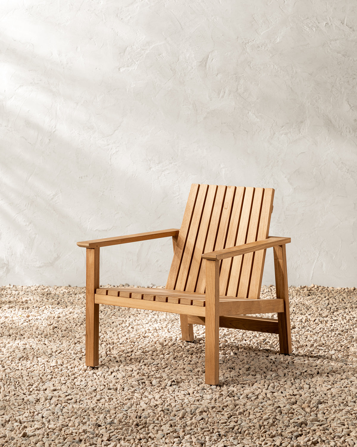 The Gilda Outdoor Lounge Chair, made of teak with a slatted back and seat, is set on gravel against a textured, light-toned wall in natural sunlight by McGee & Co.