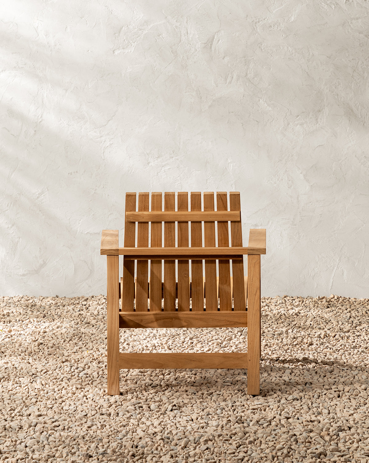 The Gilda Outdoor Lounge Chair, crafted from teak wood with vertical slats, sits on a gravel surface against a textured white wall by McGee & Co.