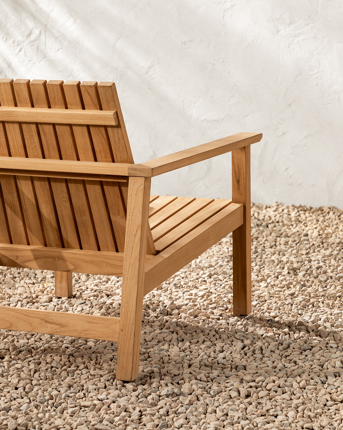 The Gilda Outdoor Lounge Chair, crafted from teak wood with a slatted back and seat, rests on small light-colored stones in front of a textured white wall, by Vendixs
