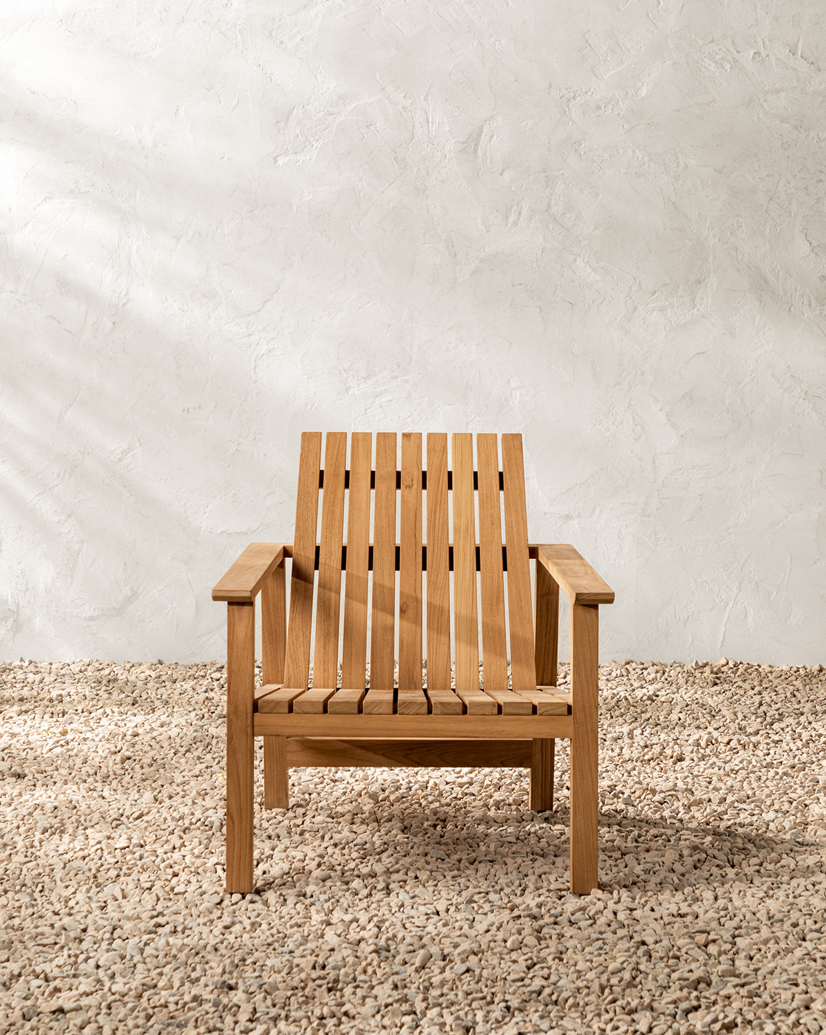 The Gilda Outdoor Lounge Chair, made of teak wood with a slatted seat and backrest, sits on gravel against a textured white wall in soft natural light, by McGee & Co.