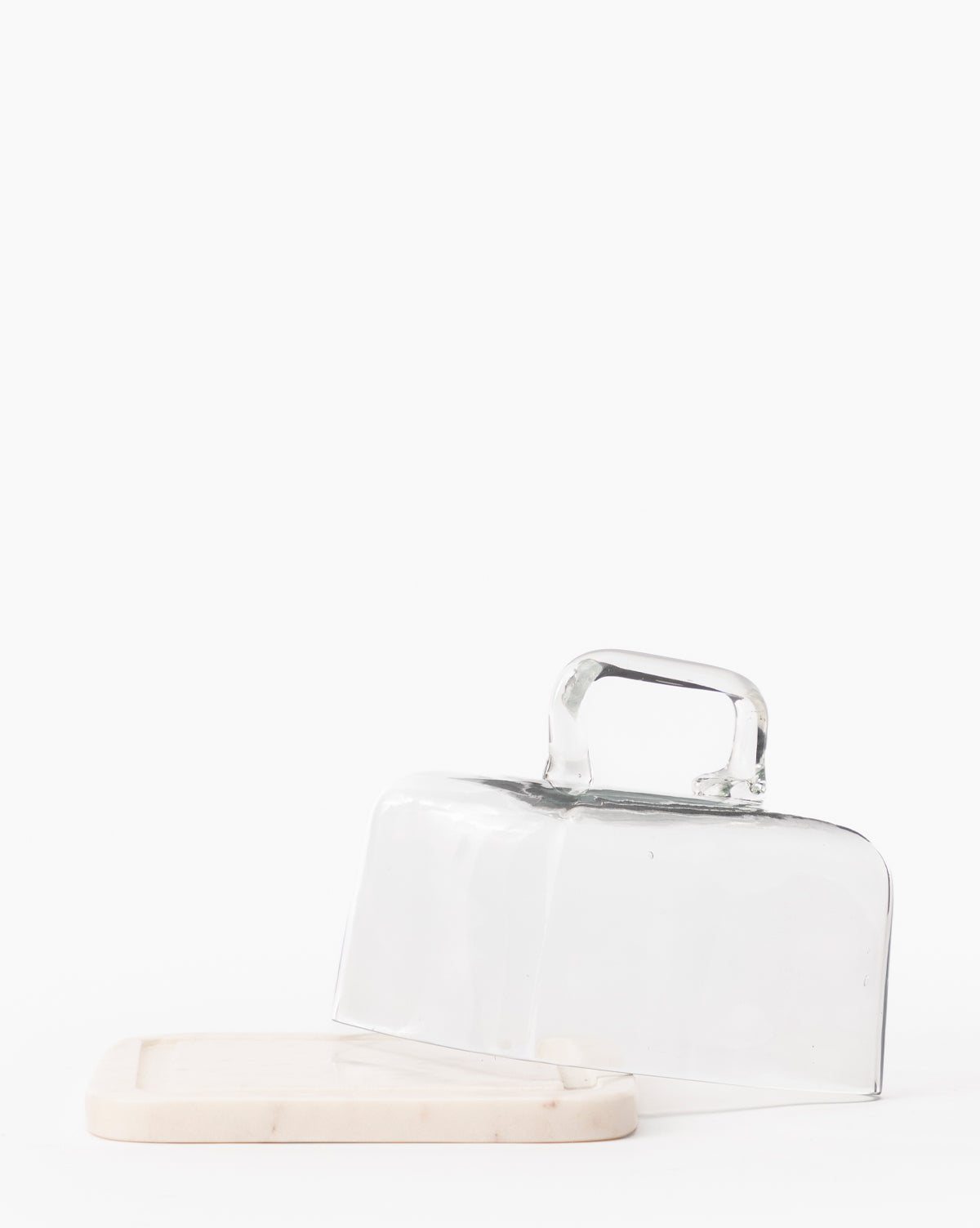The Glass & Marble Cloche features a clear glass dome with handle tilted on a sleek marble base, creating an elegant kitchen accessory set against a plain white background, by Vendixs