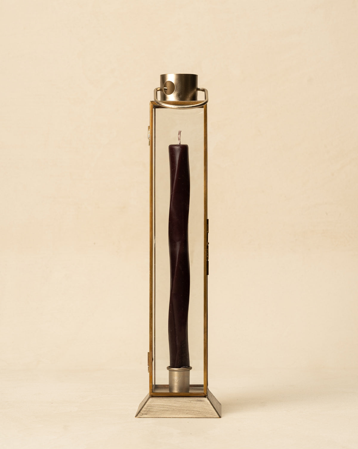The Glass & Metal Lantern Taper Holder features a dark twisted candle standing upright in its rectangular glass and metal frame with handle, creating a striking accent against a plain beige background, by Vendixs