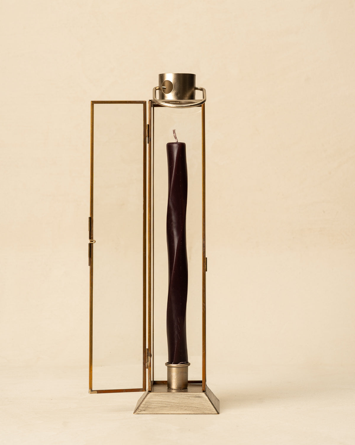 A tall, dark taper candle stands in the Glass & Metal Lantern Taper Holder with one door open, set against a plain beige background, by Vendixs