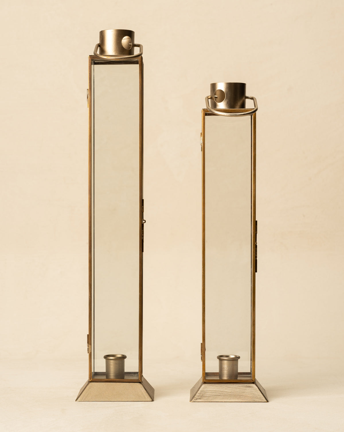 Two tall Glass & Metal Lantern Taper Holders with brass frames stand side by side on a neutral background. Each lantern has a top handle and an interior candle holder, adding stylish illumination to your space, by Vendixs
