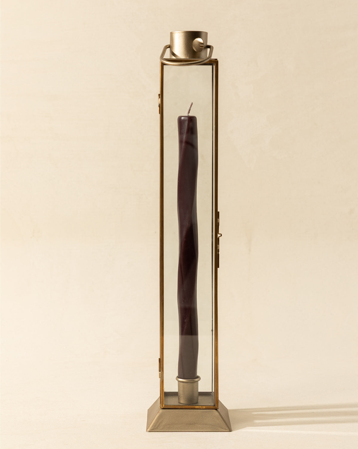 The Glass & Metal Lantern Taper Holder is a tall, rectangular lantern with clear glass sides, holding a single dark purple twisted taper candle. A small metal cup tops this elegant candle holder against a light, plain background, by Vendixs