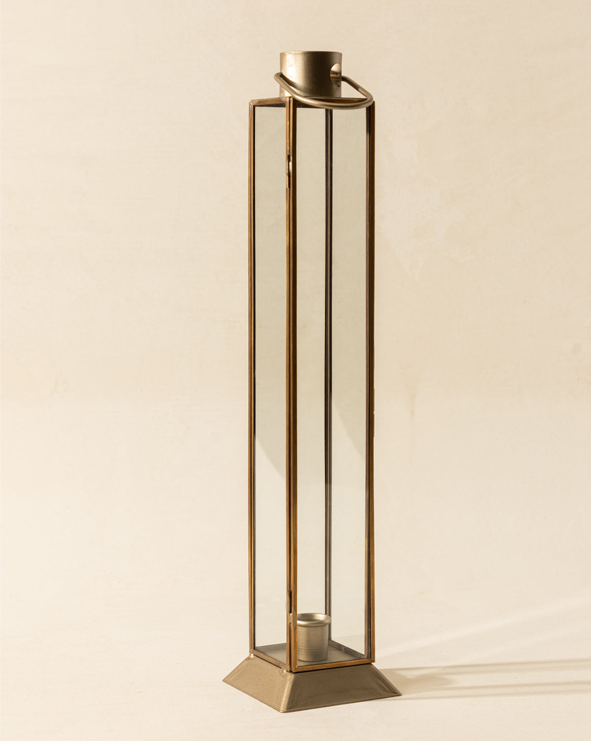 The Glass & Metal Lantern Taper Holder features a tall, rectangular design with a gold frame, wide base, and an inner round taper holder, displayed against a simple light background, by Vendixs