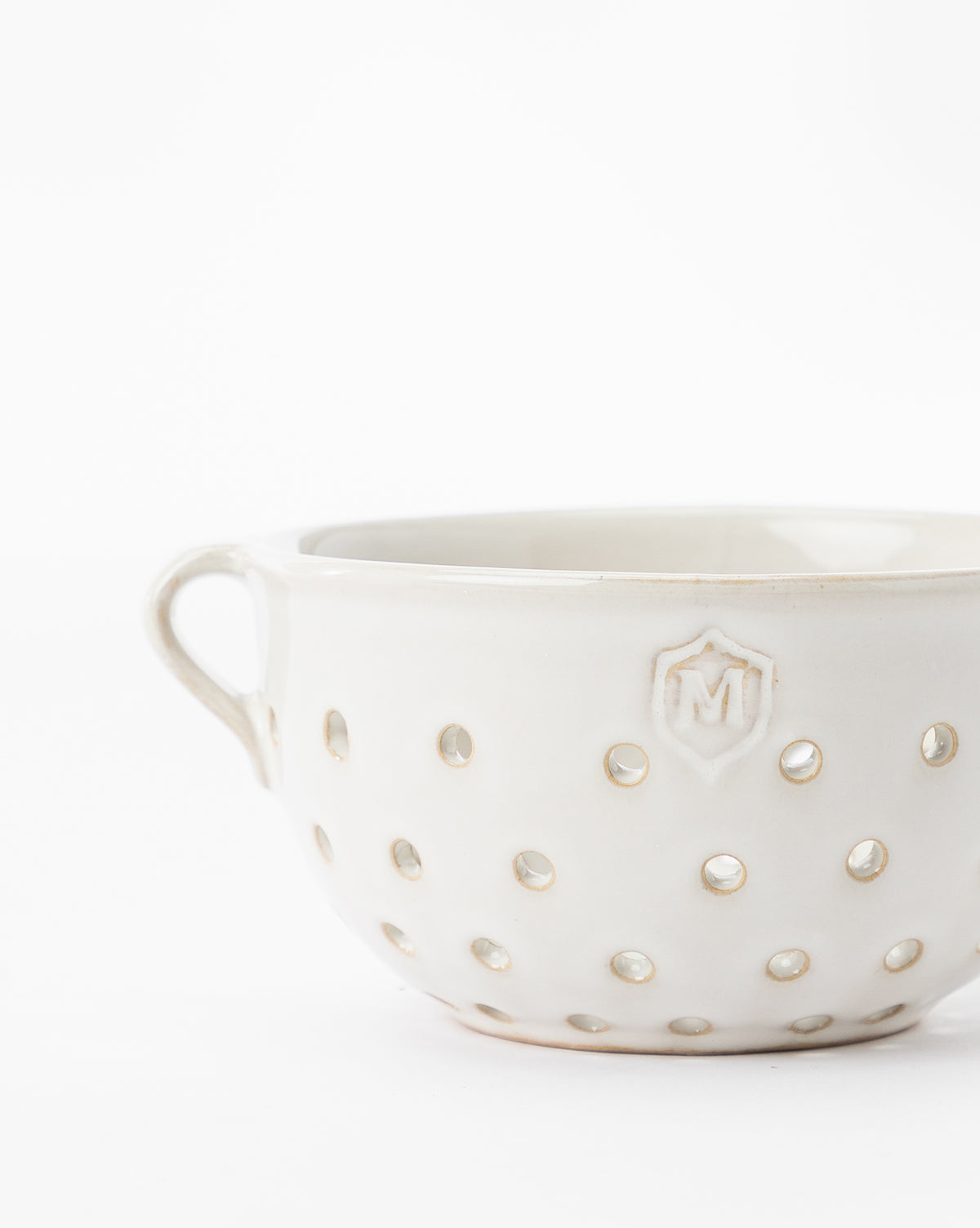 The Glazed Berry Colander is a white ceramic piece with a handle, round holes, and an embossed M emblem—perfect as both stylish serveware and elegant kitchen decor. Shown set against a plain white background, by Vendixs