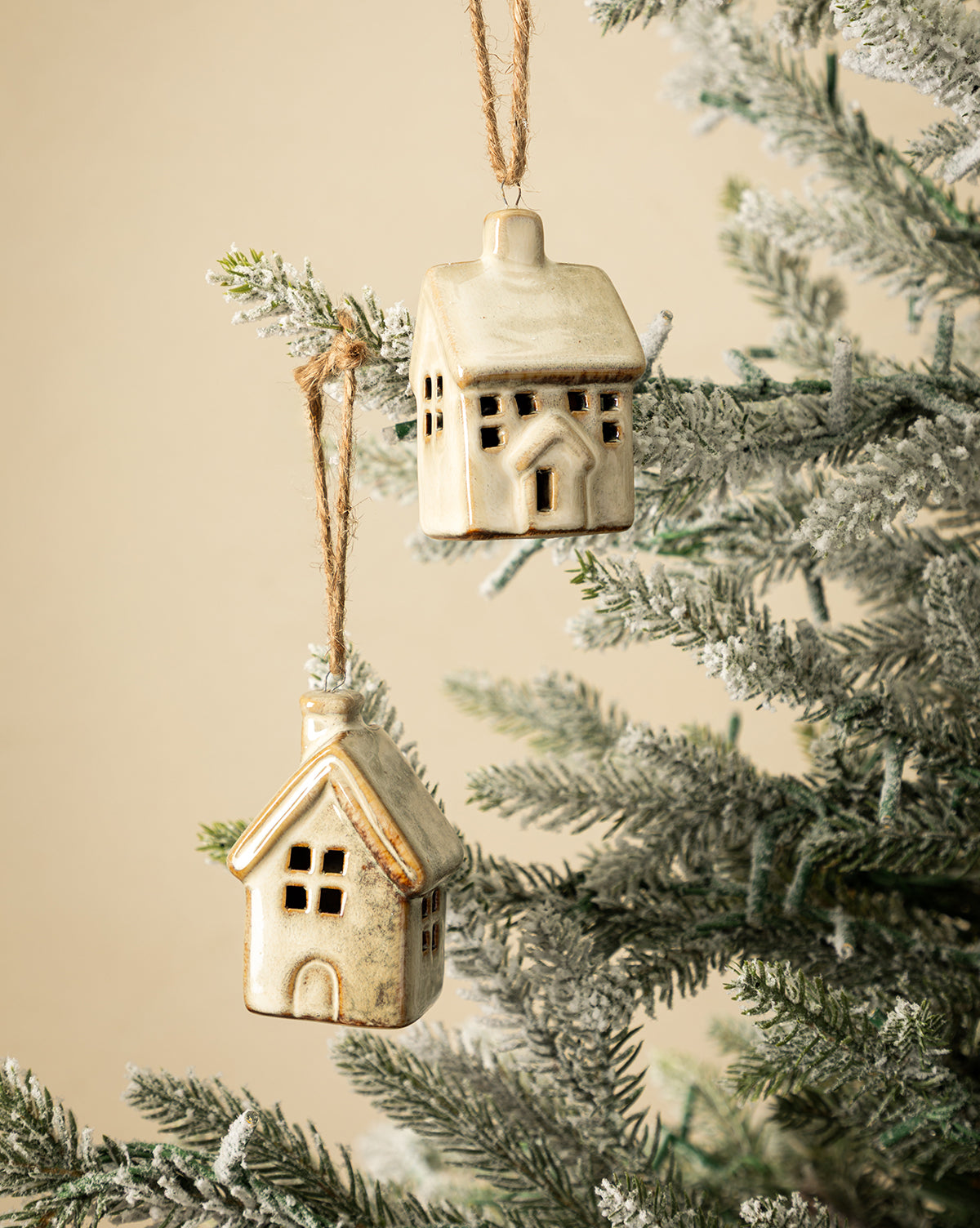 The Glazed Porcelain Houses (Set of 2) hang from twine on a snow-dusted Christmas tree branch against a beige background, bringing charming festive decor to your holiday setting, by Vendixs