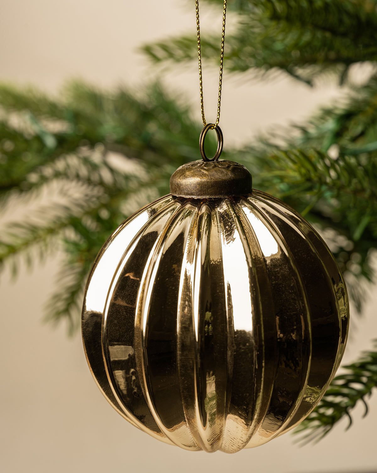 The 3" Gold Bauble, a shiny glass Christmas ornament with a gold finish, hangs from a gold string on a green pine branch against a soft beige background, by Vendixs