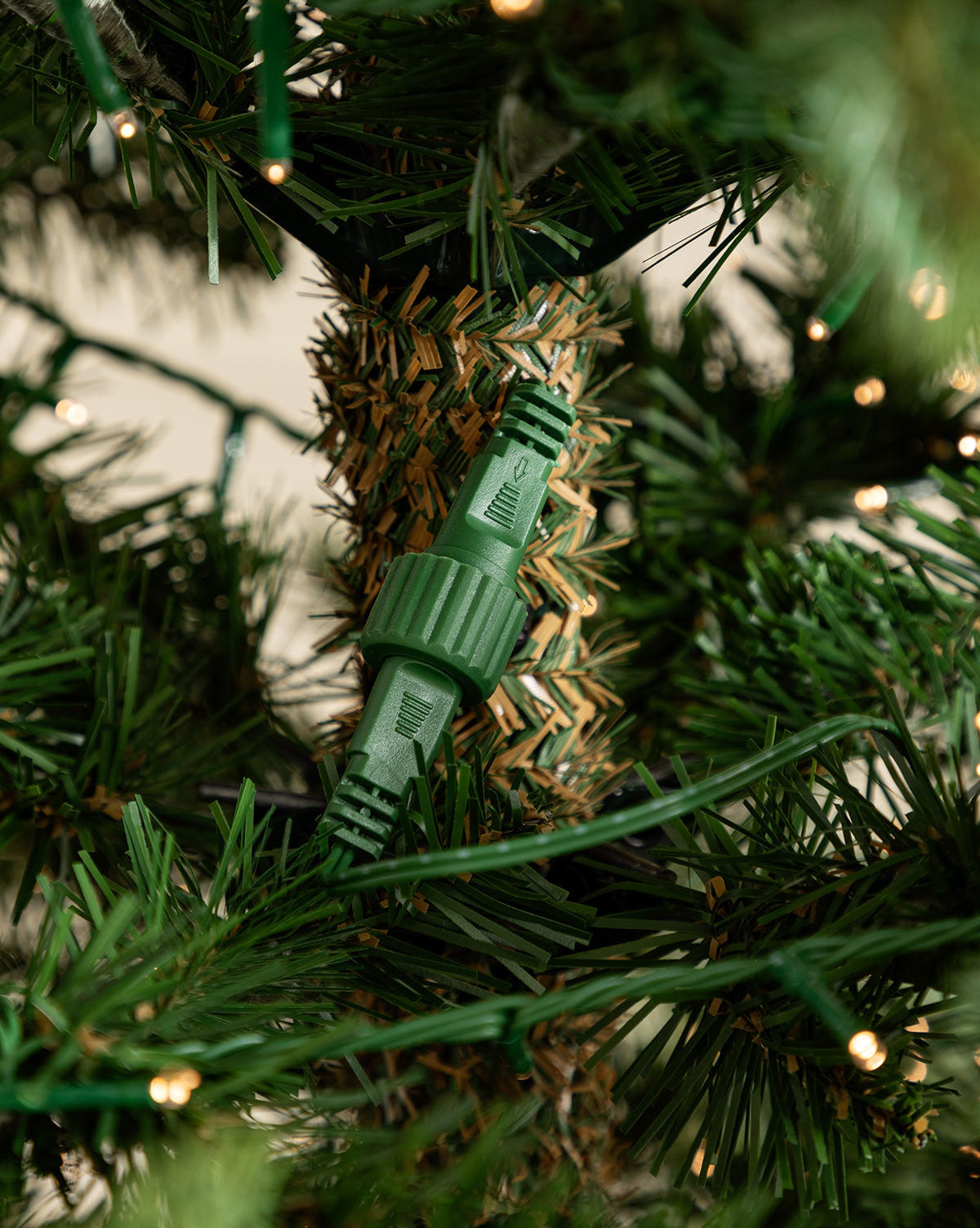 A green electrical connector plugs into Christmas lights among the lush branches of the 7' Grandis Fir Pre-Lit Faux Tree, by Vendixs