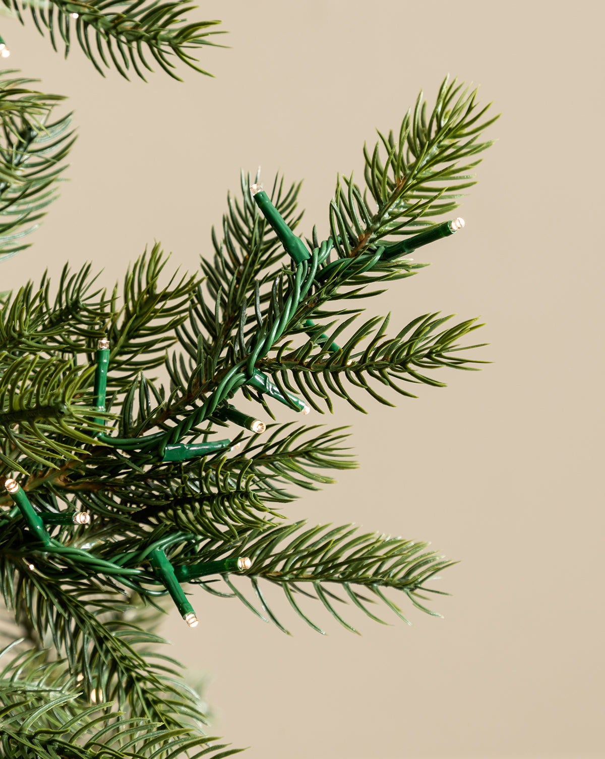 A close-up of green branches from the 7' Grandis Fir Pre-Lit Faux Tree, featuring small white string lights, set against a plain beige background, by Vendixs