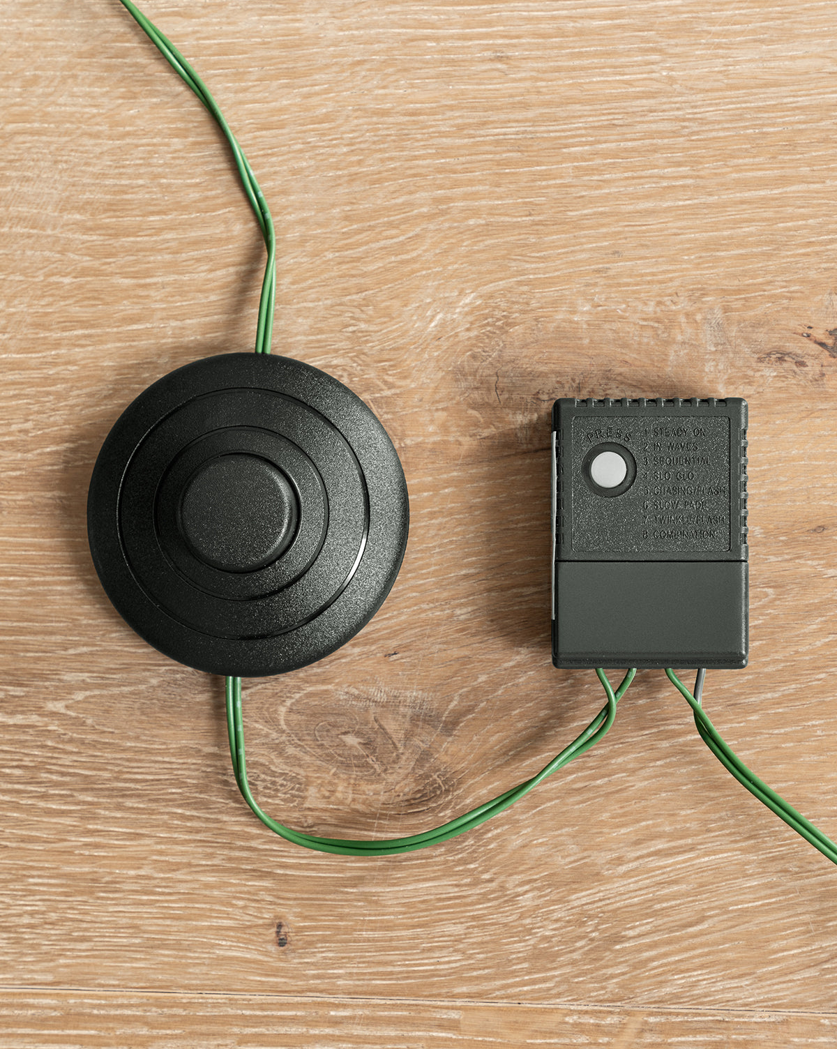 A black circular foot pedal, wired to a small black control box, connects to the 7' Grandis Fir Pre-Lit Faux Tree; both devices rest on a light wood surface, by Vendixs