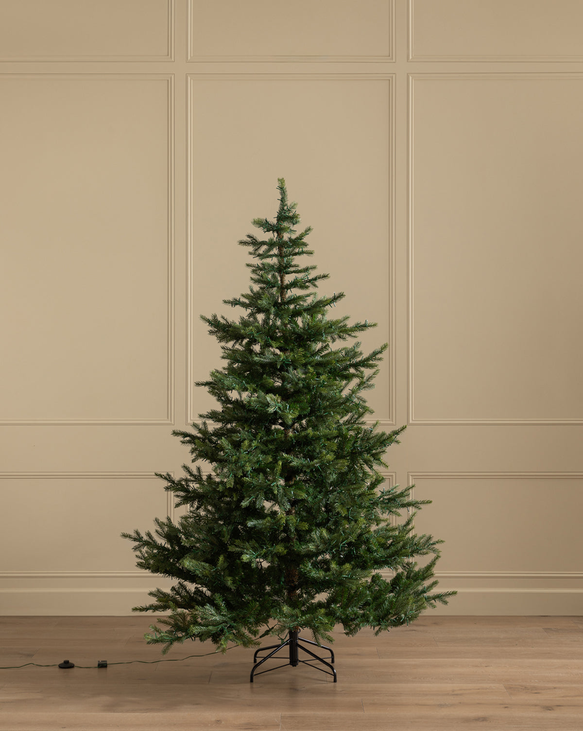 A 7' Grandis Fir Pre-Lit Faux Tree with green branches stands upright on a metal stand in a room with beige paneled walls and a wooden floor, with an electrical cord lying on the floor to the left of the tree, by Vendixs