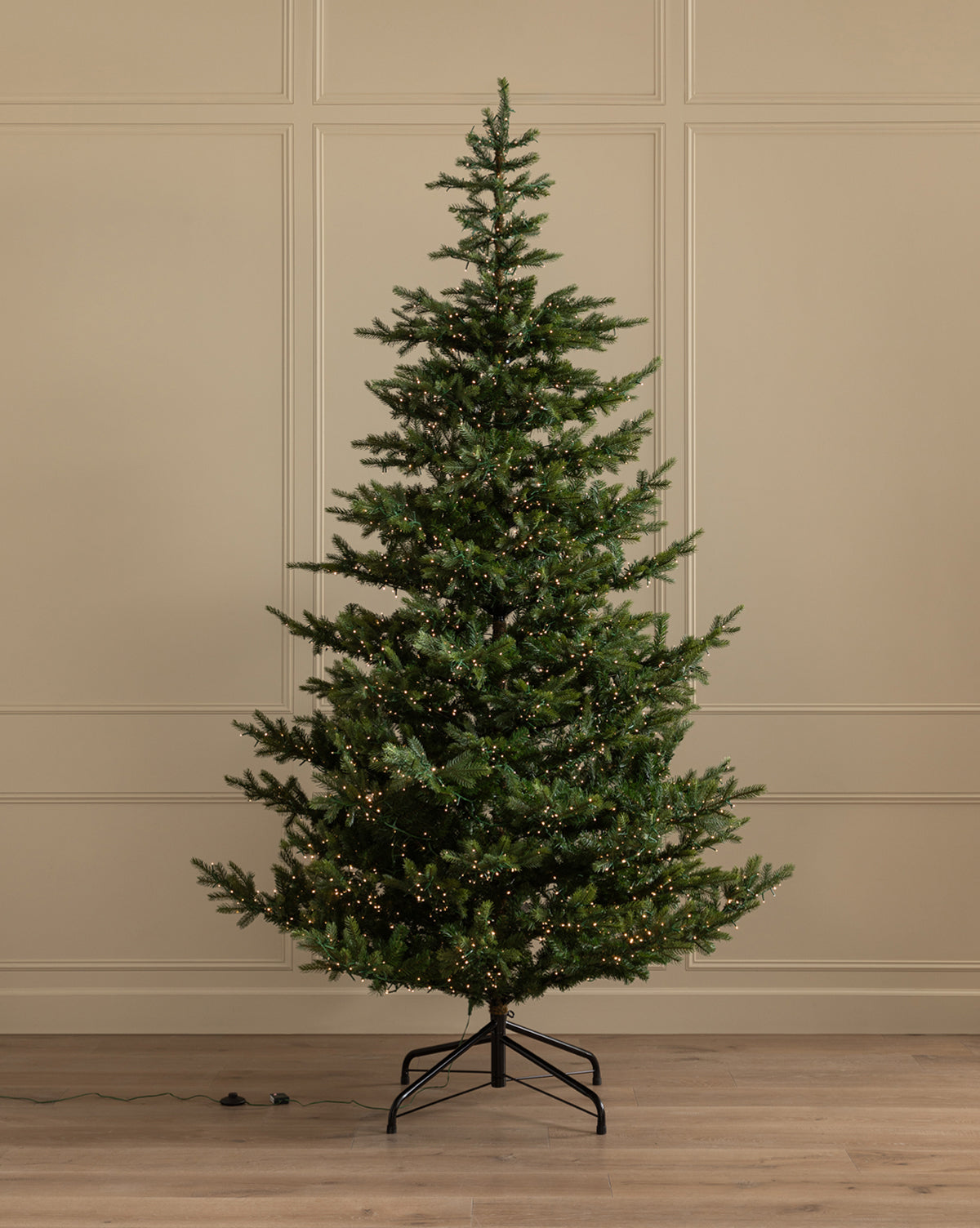 The 9' Grandis Fir Pre-Lit Faux Tree, with sparse green branches and small white lights, stands on a metal base in a room with light walls and wooden floor, by Vendixs