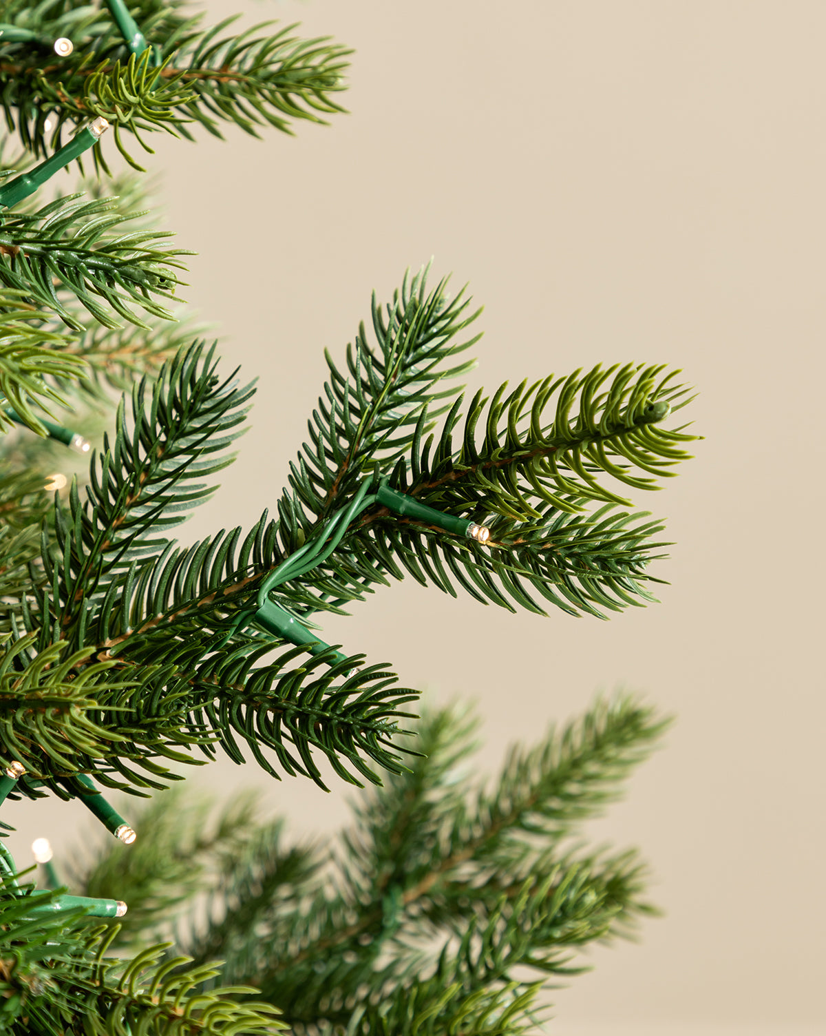 Close-up of 9' Grandis Fir Pre-Lit Faux Tree branches decorated with small white string lights, casting a festive glow against a plain beige background, by Vendixs