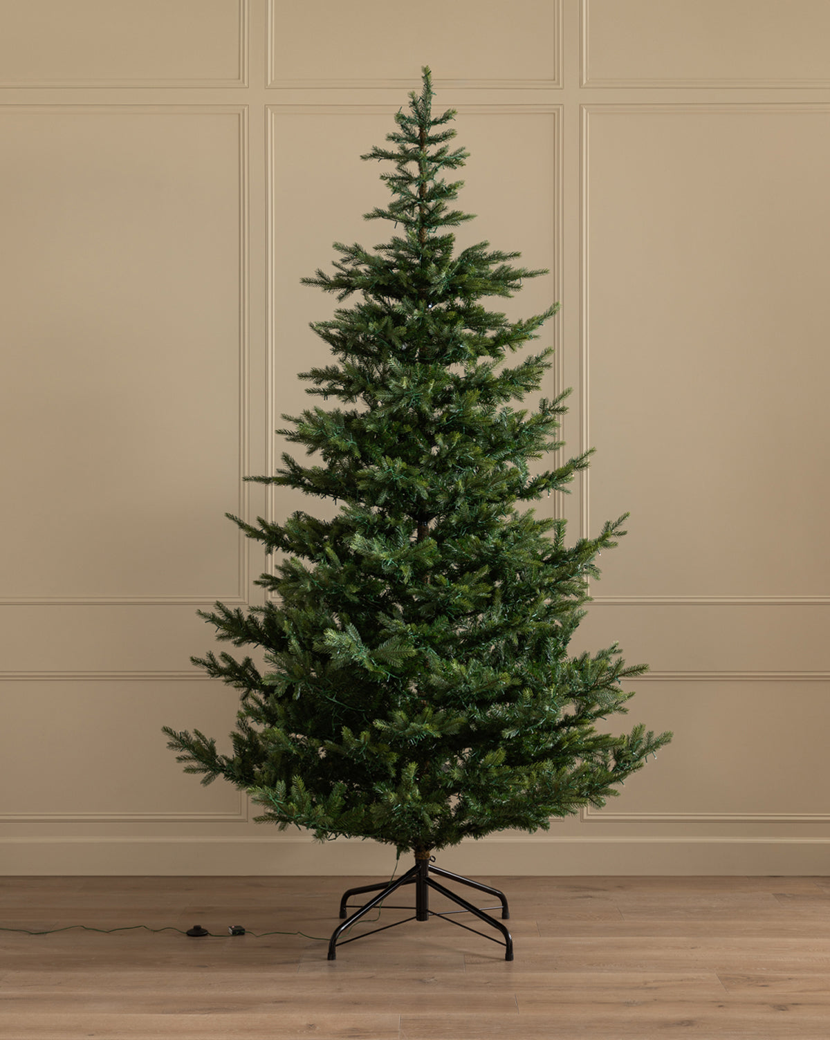 The 9' Grandis Fir Pre-Lit Faux Tree stands on a metal base in a room with beige paneled walls and a wooden floor, appearing undecorated and unlit, with its plug and wire visible on the floor, by Vendixs