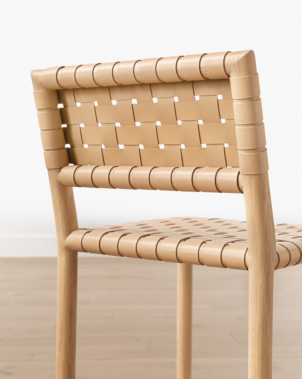 A close-up view of the Greely Stool, featuring a white oak frame with woven aniline-dyed leather backrest and seat, set on a light wood floor against a white wall, by Vendixs