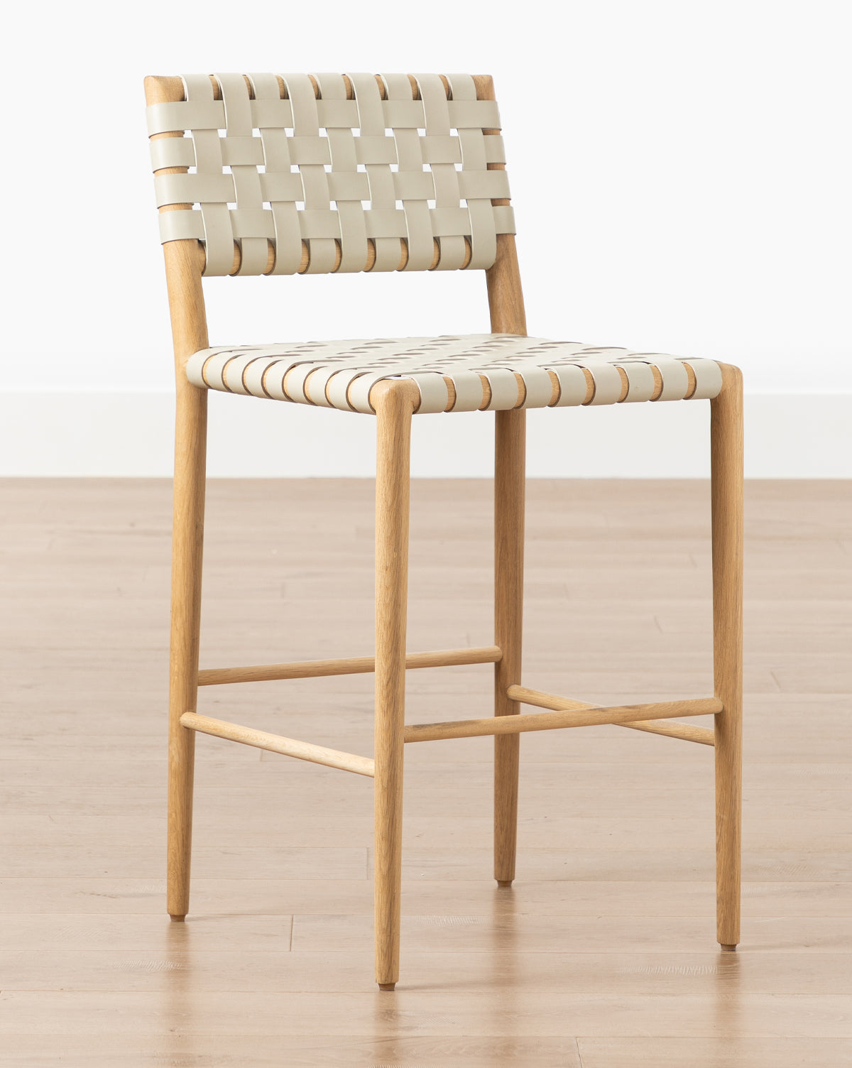 The Greely Stool features a modern white oak frame with beige woven straps for the seat and backrest, shown on a light wood floor against a plain white wall by McGee & Co.