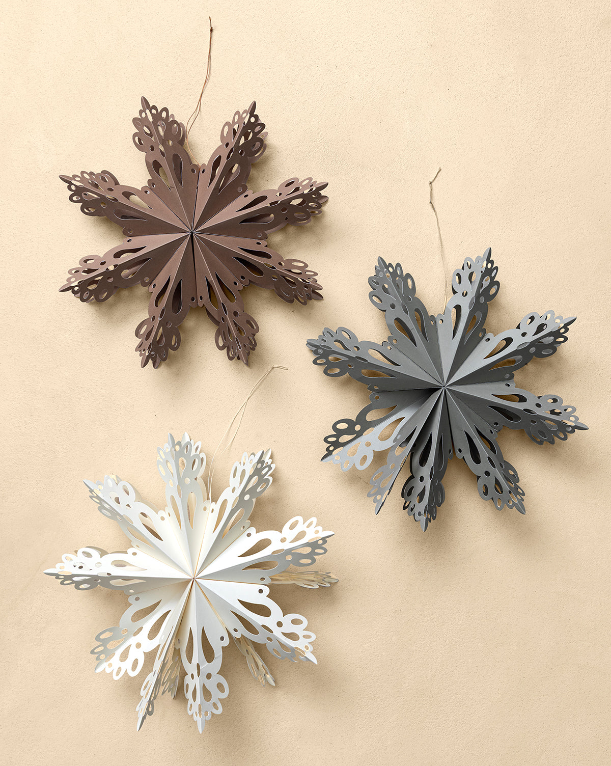 The Handmade Assorted Recycled Paper Snowflakes (Set of 3) features ornaments in brown, gray, and white with intricate cut-out details and strings for easy holiday decorating, shown hanging against a plain beige background, by Vendixs