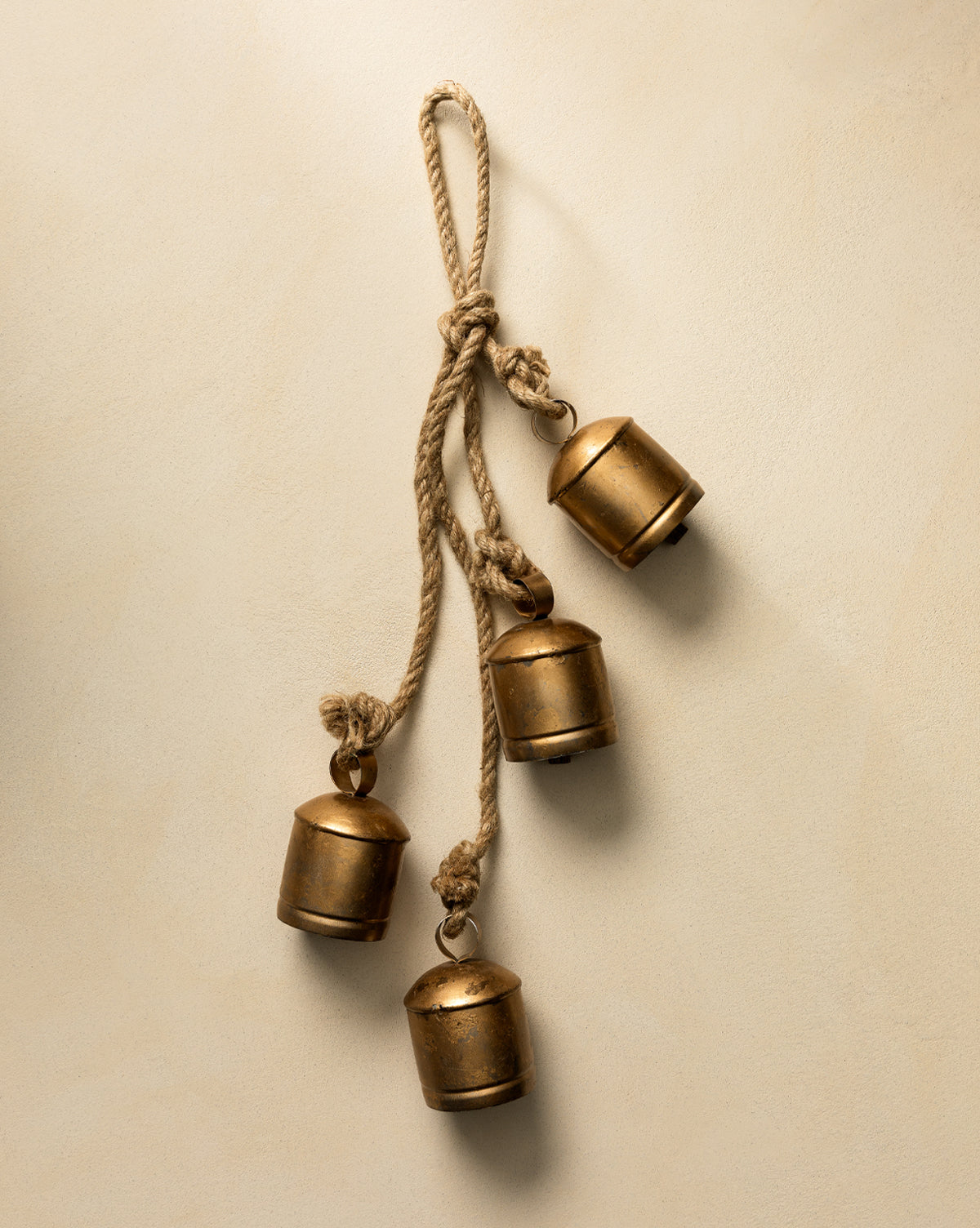 The Hanging Iron Bells set features four small, gold-finished bells with rounded tops hanging at varying lengths from a twisted rope—ideal for holiday décor against a neutral backdrop, by Vendixs