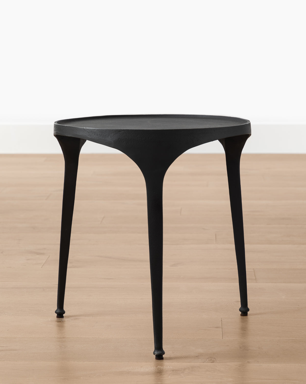 The Haskell Side Table, small and round with three slender legs, stands on a light wood floor by a plain white wall, highlighting its sleek contemporary design, by Vendixs