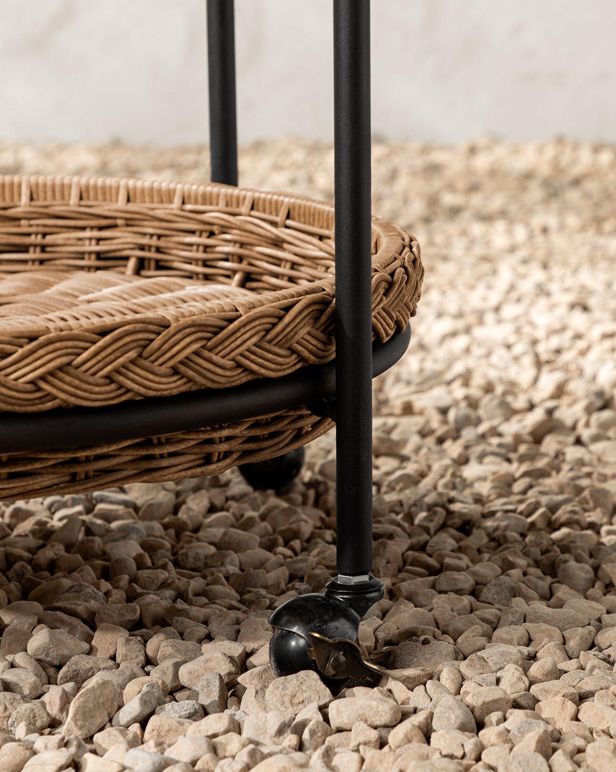 A close-up of the Haviland Bar Cart featuring weather-resistant wicker, a black metal frame, and caster wheels, set on a bed of small, light-colored pebbles, by Vendixs