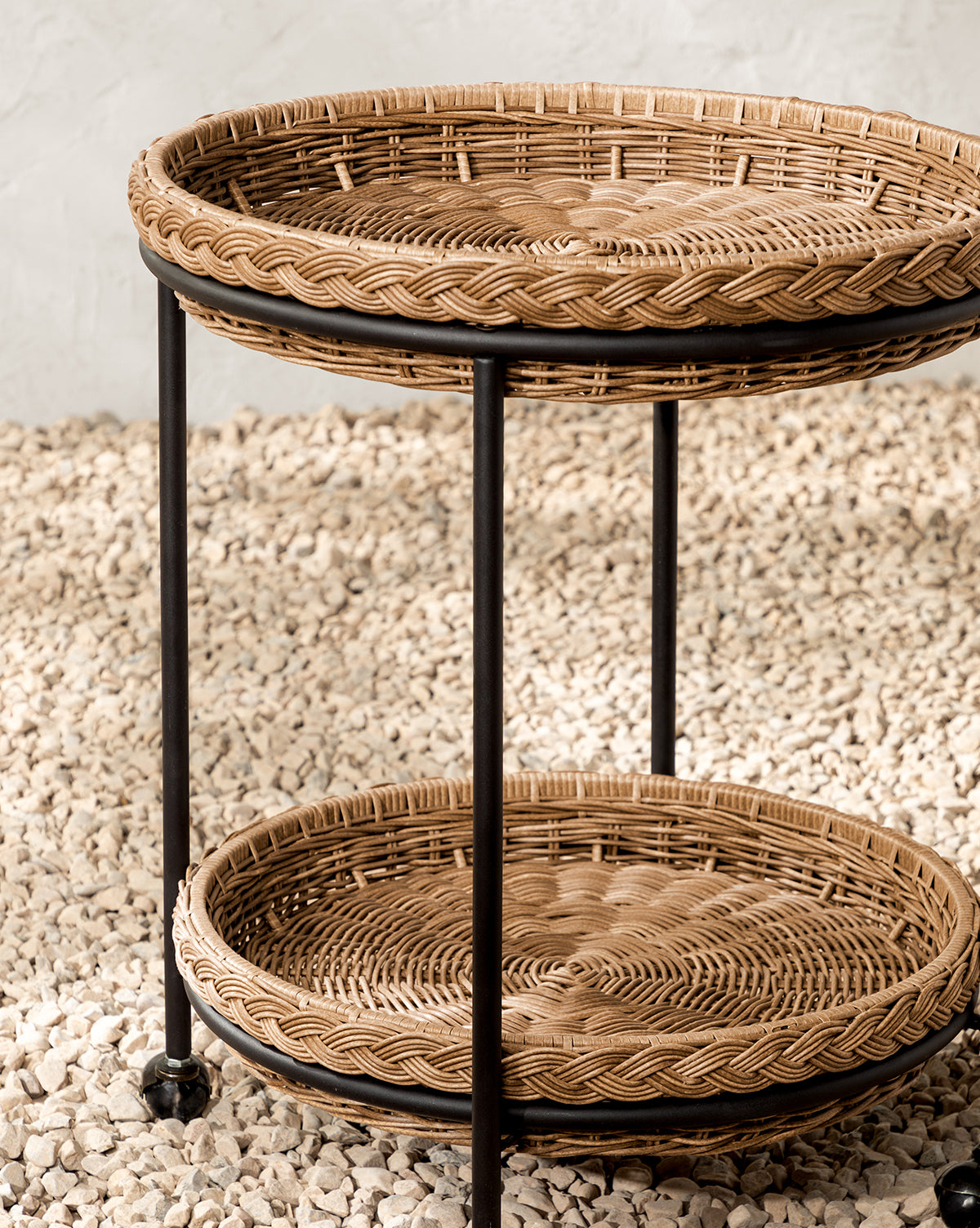 The Haviland Bar Cart features weather-resistant wicker with two round tiers and sturdy black metal legs, set on small white stones against a light textured wall—ideal for outdoor entertaining, by Vendixs