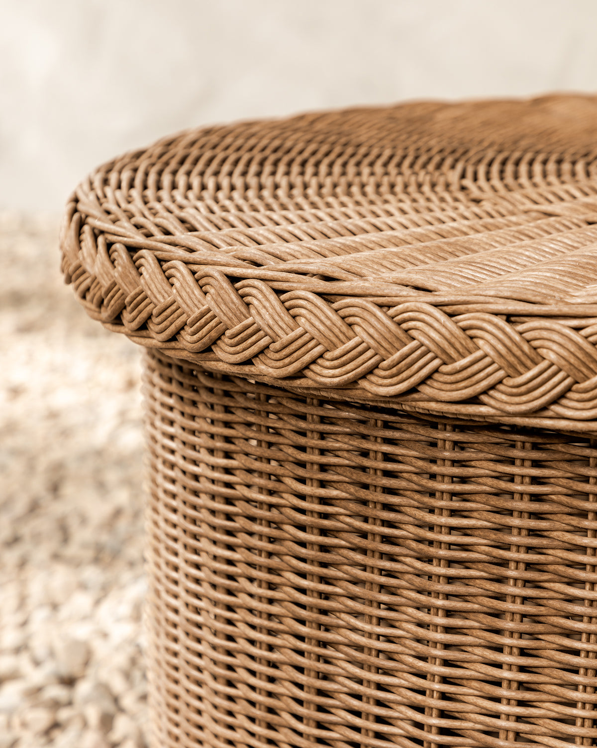Close-up of the Haviland Coffee Table, a round wicker piece with a braided rim and intricate weaving. This weather-resistant table sits outdoors on small pebbles, by Vendixs