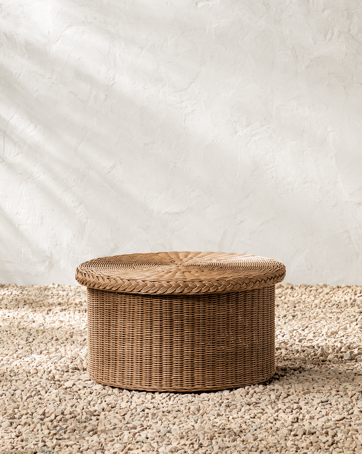 The Haviland Coffee Table, featuring a weather-resistant wicker woven design, sits on small pebbles by a textured white wall as soft natural light casts gentle shadows by McGee & Co.