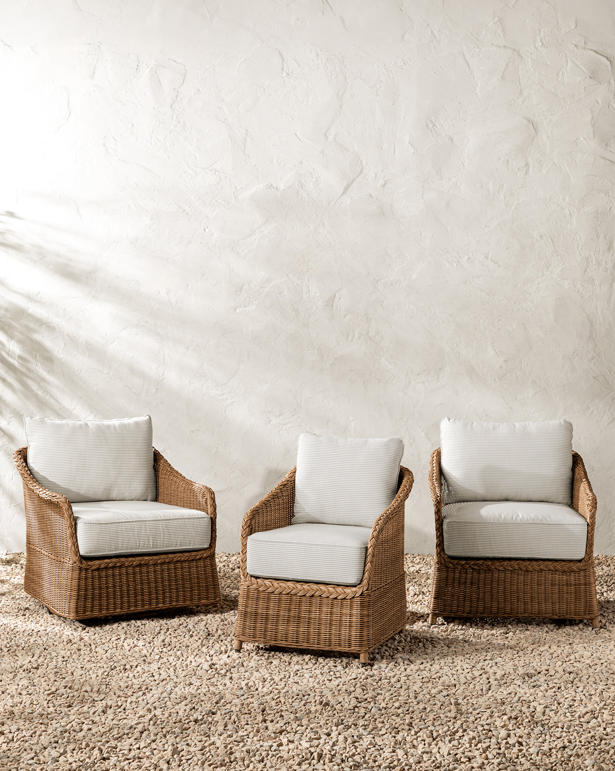 Three Haviland Outdoor Lounge Chairs with Perennials Taupe Stripe cushions are arranged on a pebble floor against a textured white wall. Soft natural light casts gentle shadows, creating a calm and inviting atmosphere by McGee & Co.
