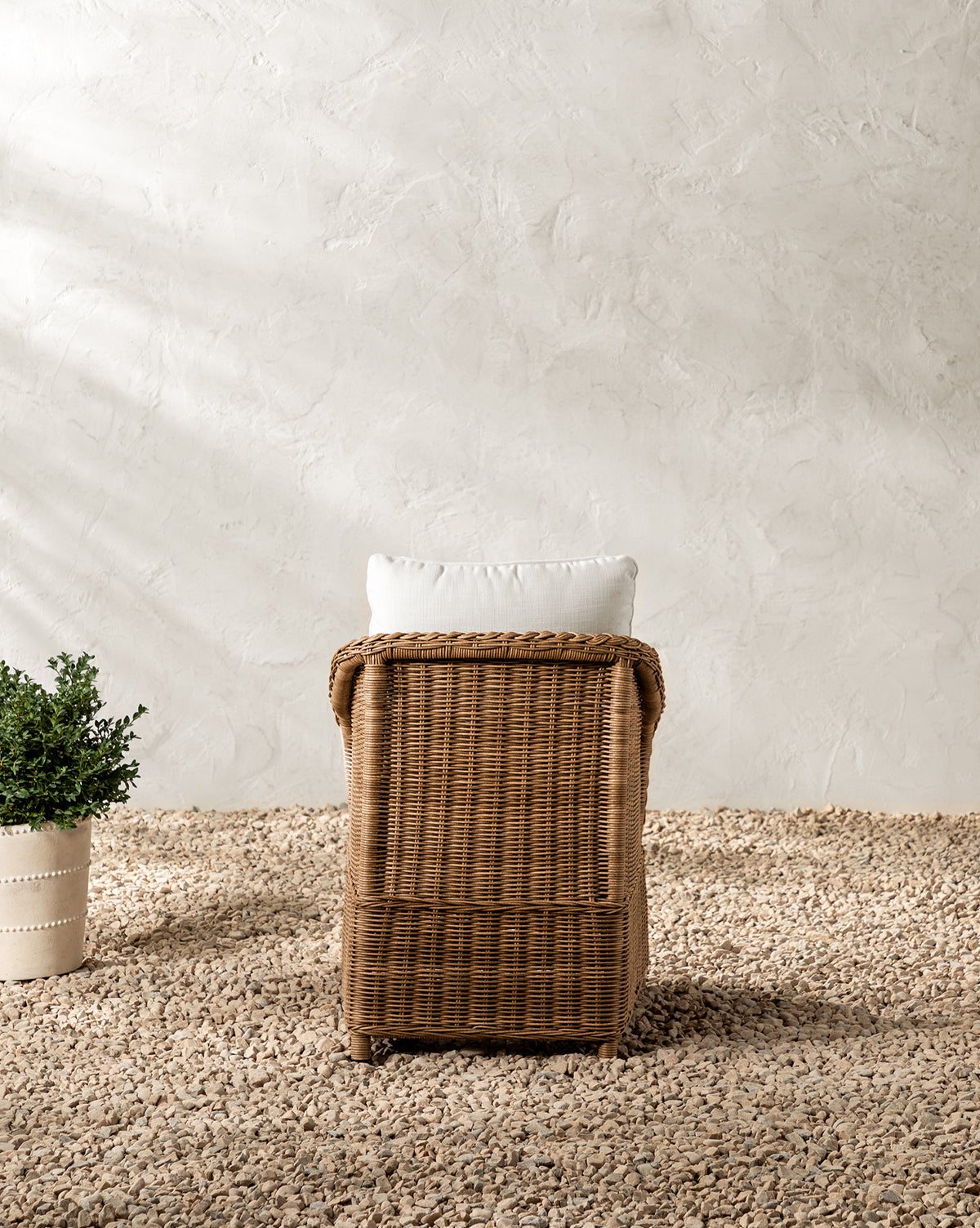 The Haviland Outdoor Dining Chair with a white cushion sits on a pebble-covered floor. To the left is a potted green plant, all set against a textured, light-colored wall in the background, by Vendixs
