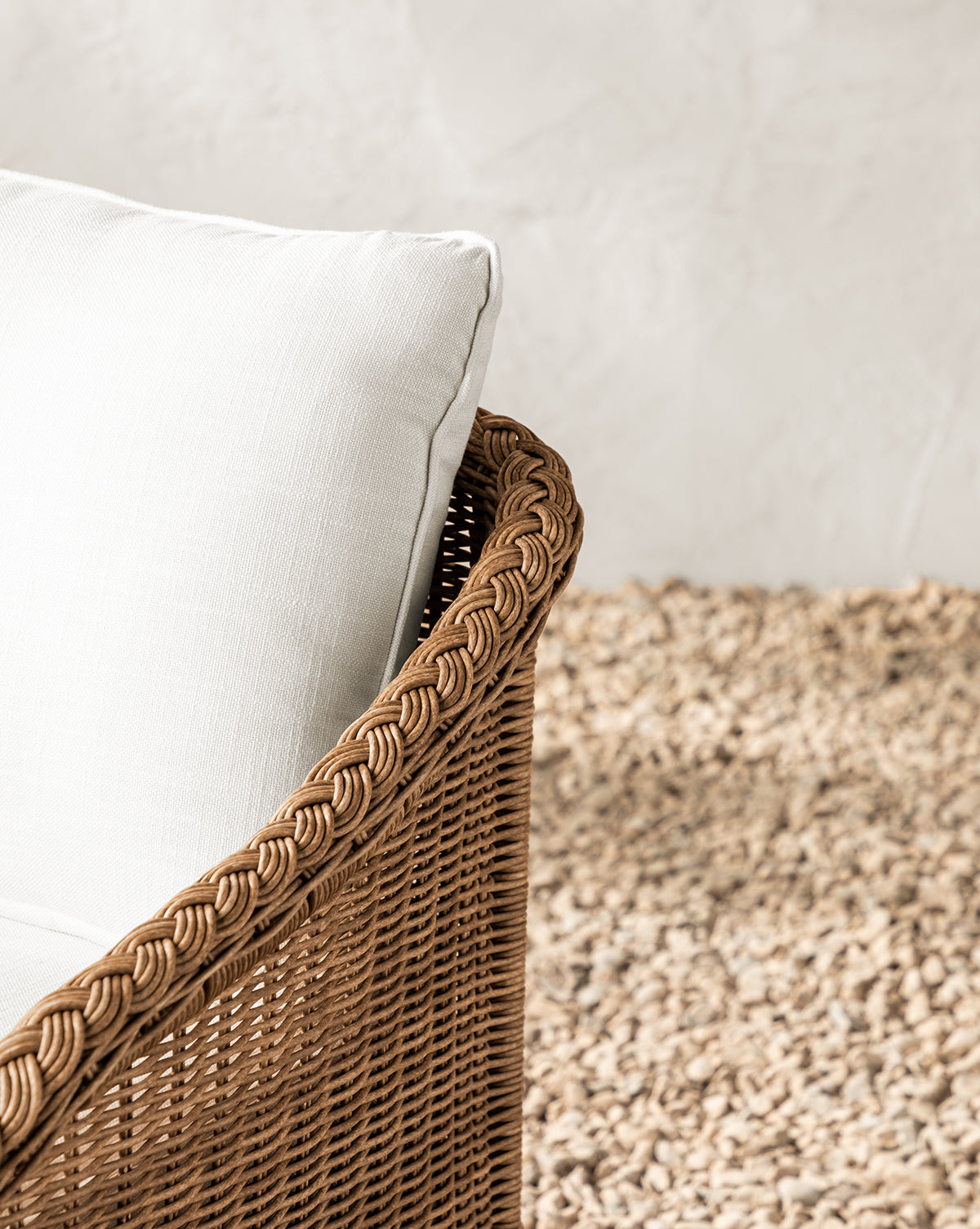 The Haviland Outdoor Dining Chair, crafted from weather-resistant wicker with a white cushion, sits atop a textured beige rug. Its intricate woven design stands out against a light, textured background wall, by Vendixs