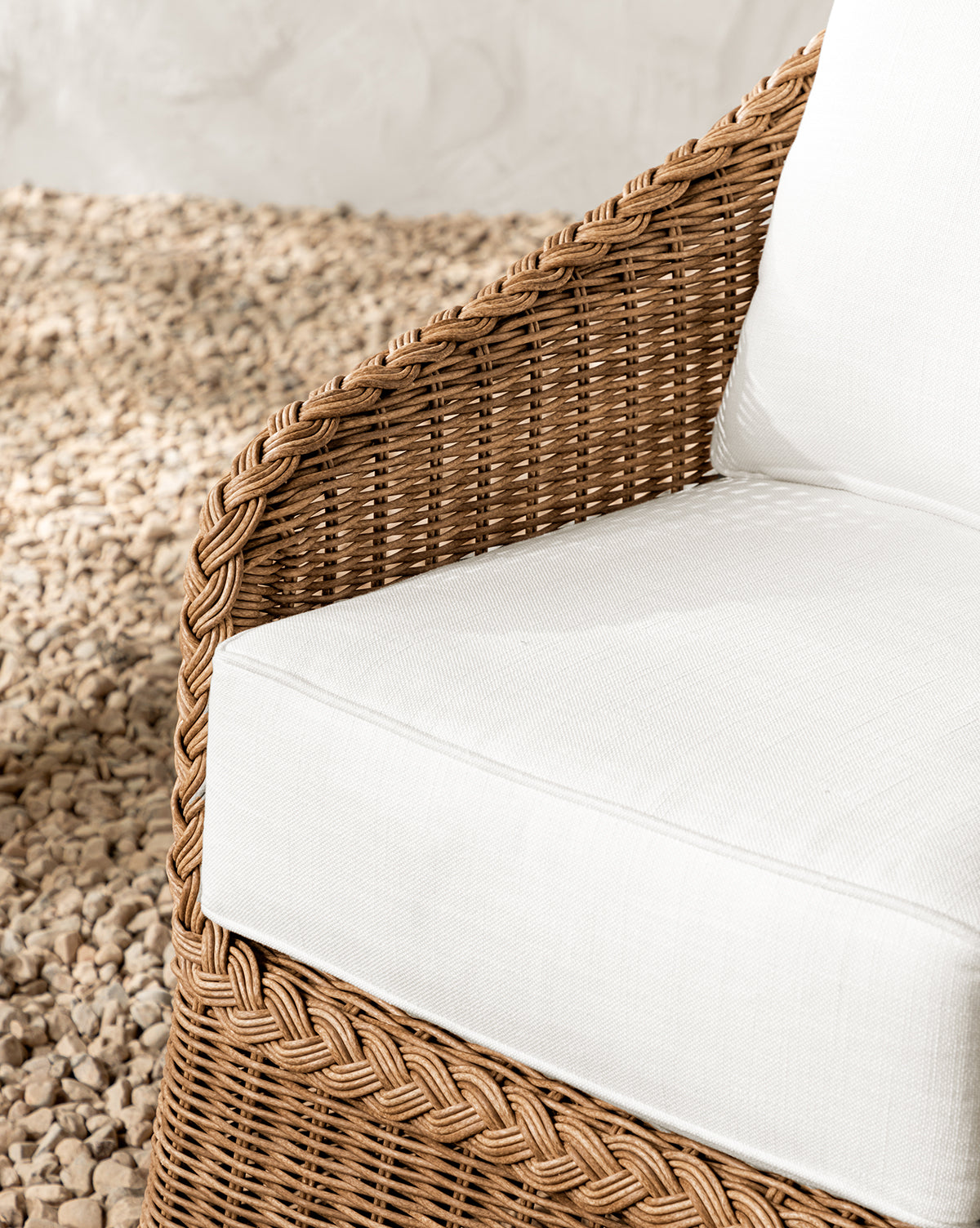 Close-up of the Haviland Outdoor Dining Chair, weather-resistant with a white cushion and pillow, set on light stones. Features include a braided wicker armrest and textured backrest, making it ideal for stylish outdoor dining, by Vendixs