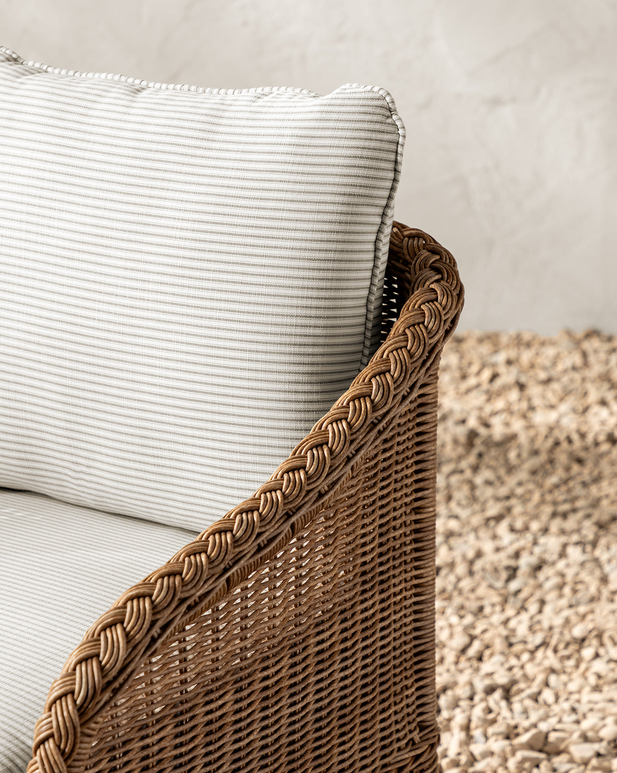 Close-up of the Haviland Outdoor Dining Chair with Perennials Taupe Stripe cushions, featuring a weather-resistant wicker frame, braided armrest detail, and set on light-colored stones before a textured wall, by Vendixs