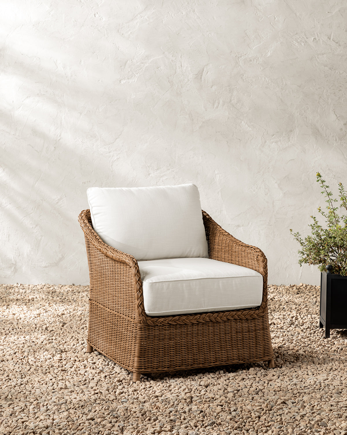 The Haviland Outdoor Lounge Chair, featuring weather-resistant wicker and white cushions, is placed on small pebbles before a textured white wall, beside a black planter filled with lush green foliage, by McGee & Co.