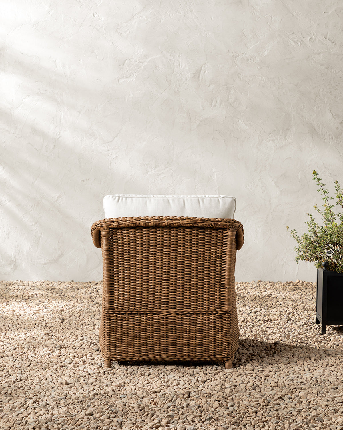 The Haviland Outdoor Lounge Chair, featuring a white cushion and weather-resistant wicker, is set on gravel before a textured white wall, accompanied by a potted plant on the right, by McGee & Co.