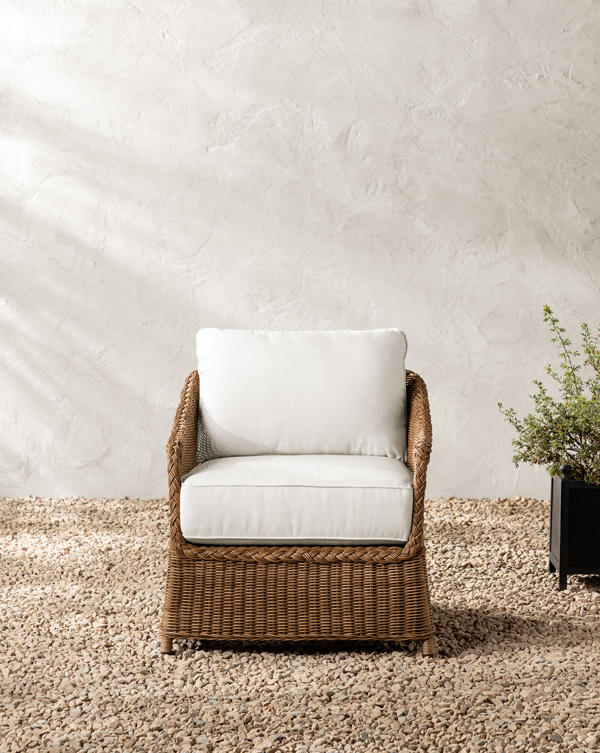 The Haviland Outdoor Lounge Chair with weather-resistant wicker and white cushions sits on gravel before a textured, light wall. To its right, a black rectangular planter filled with green foliage completes the scene, by Vendixs