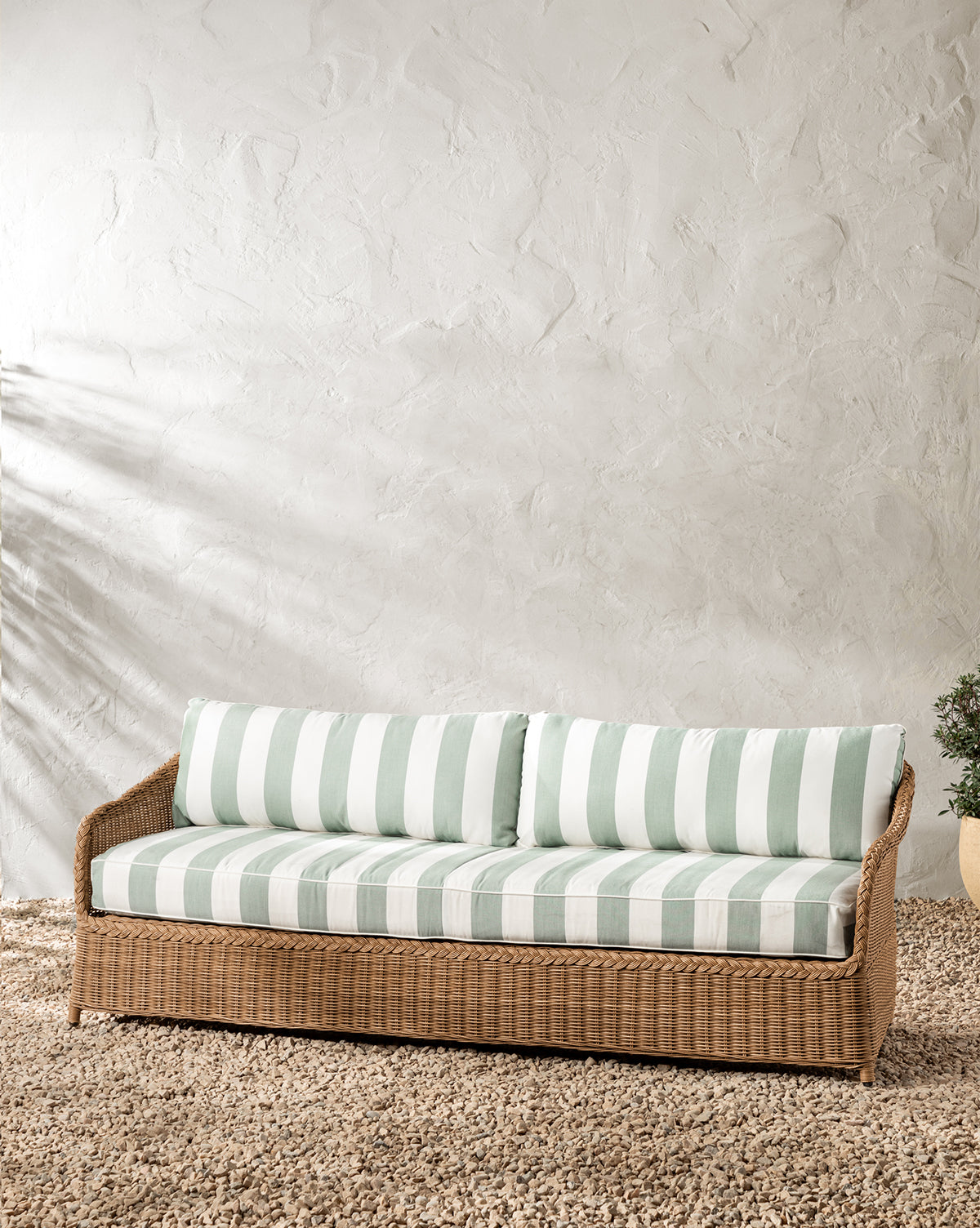 The Haviland Outdoor Sofa with Striped Cushions 88 sits on a textured beige rug against a white stucco wall for an inviting setup, with a small green plant visible on the right, by McGee & Co.