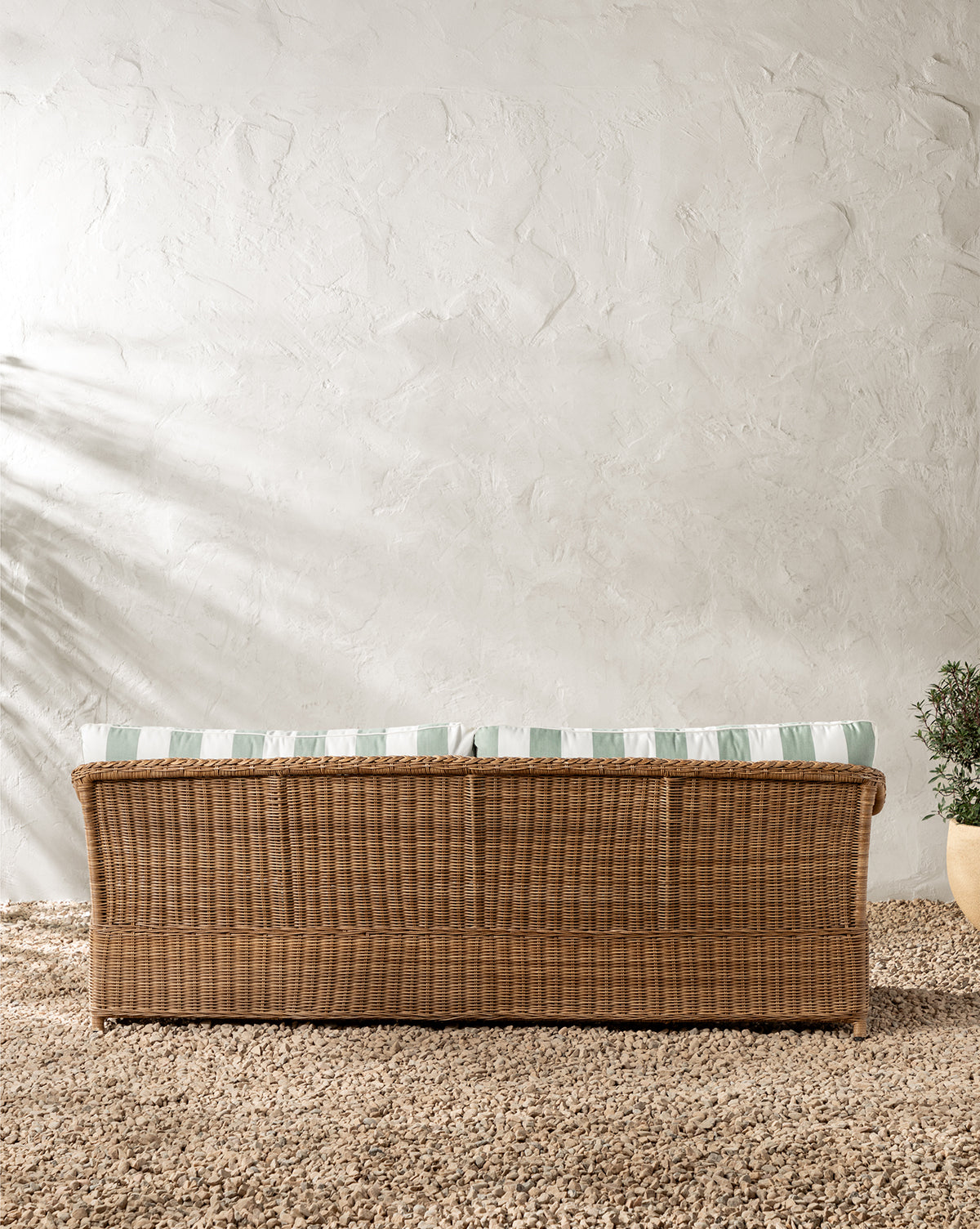 The Haviland Outdoor Sofa with Striped Cushions 88 sits on a pebble floor against a textured white wall, while a potted plant to the right and soft shadows complete this tranquil outdoor setting, by McGee & Co.