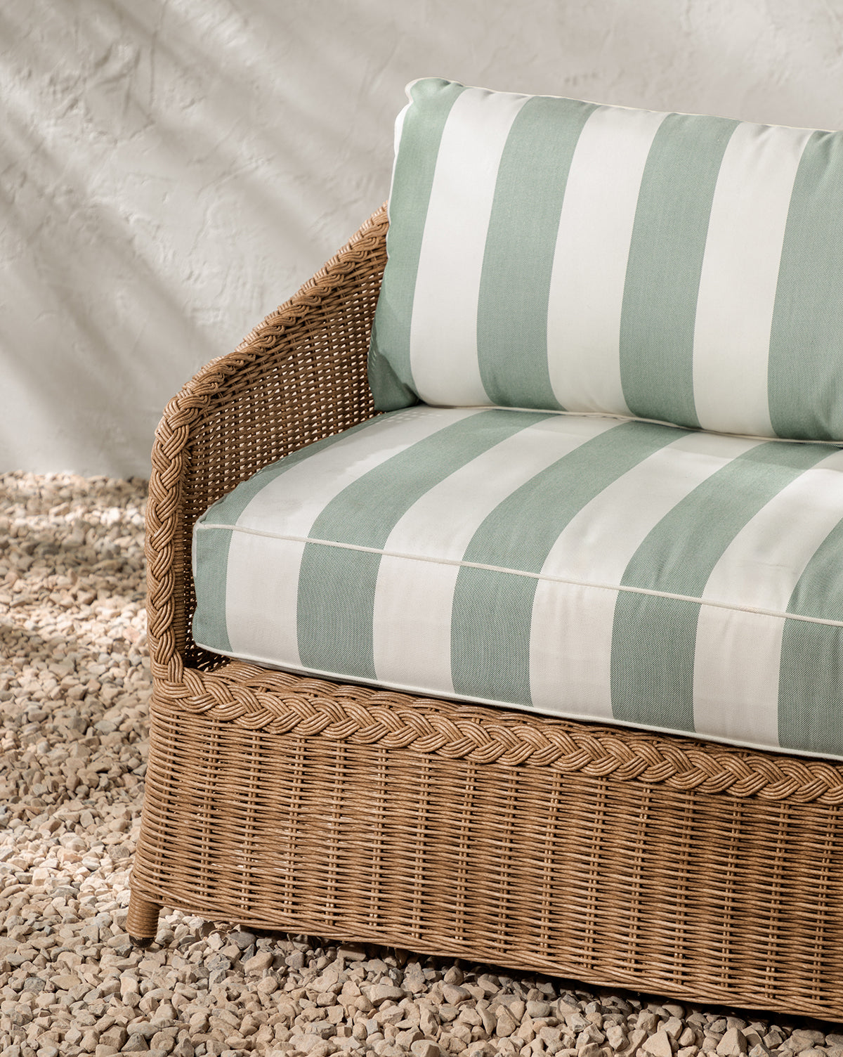 The Haviland Outdoor Sofa with Striped Cushions 88 rests on small white pebbles against a textured white wall, while sunlight casts soft shadows over its green and white cushions, by Vendixs