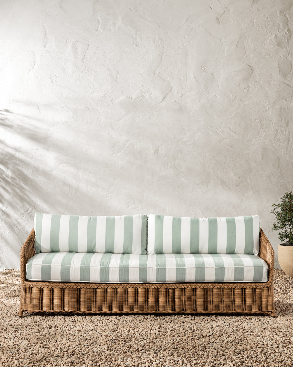 The Haviland Outdoor Sofa with Striped Cushions 88 rests on a gravel floor against a textured white wall, featuring light green and white stripes. A small potted plant sits to the right as leafy shadows fall gently across the scene, by McGee & Co.